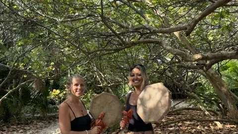 Karina and Alexandra holding frame drums on the sand under a sea grape tree