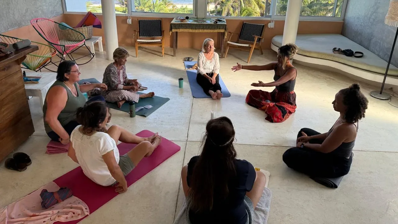Karina guiding a group of women seated on yoga mats in a circle, the sea visible through tall windows