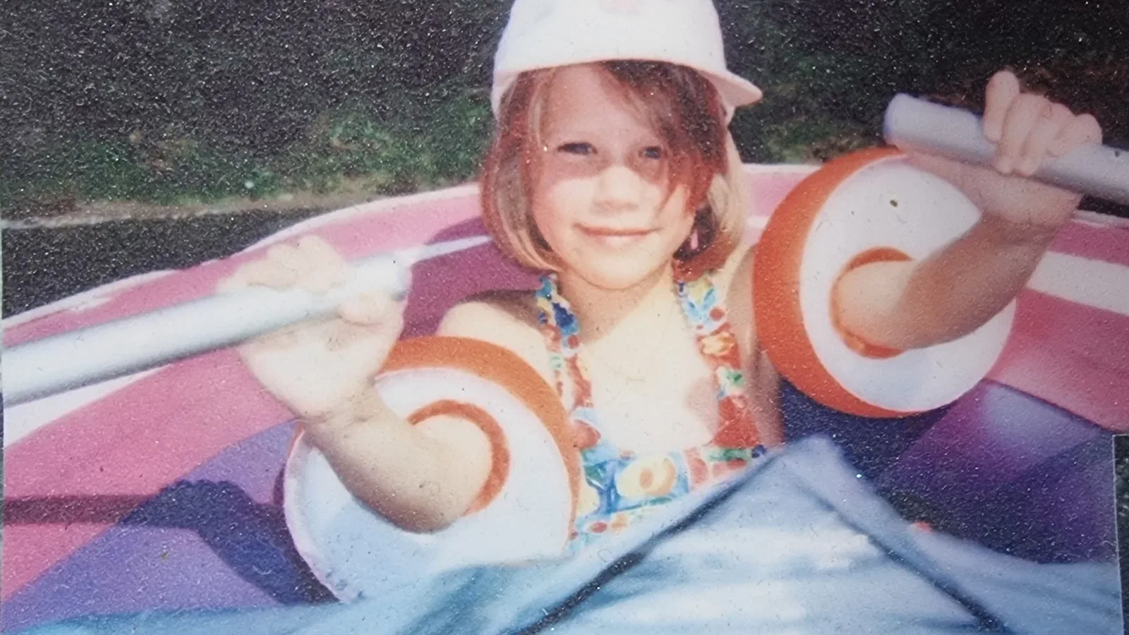 Alexandra as a little girl in a sun hat and arm floats, paddling an inflatable boat, 1989