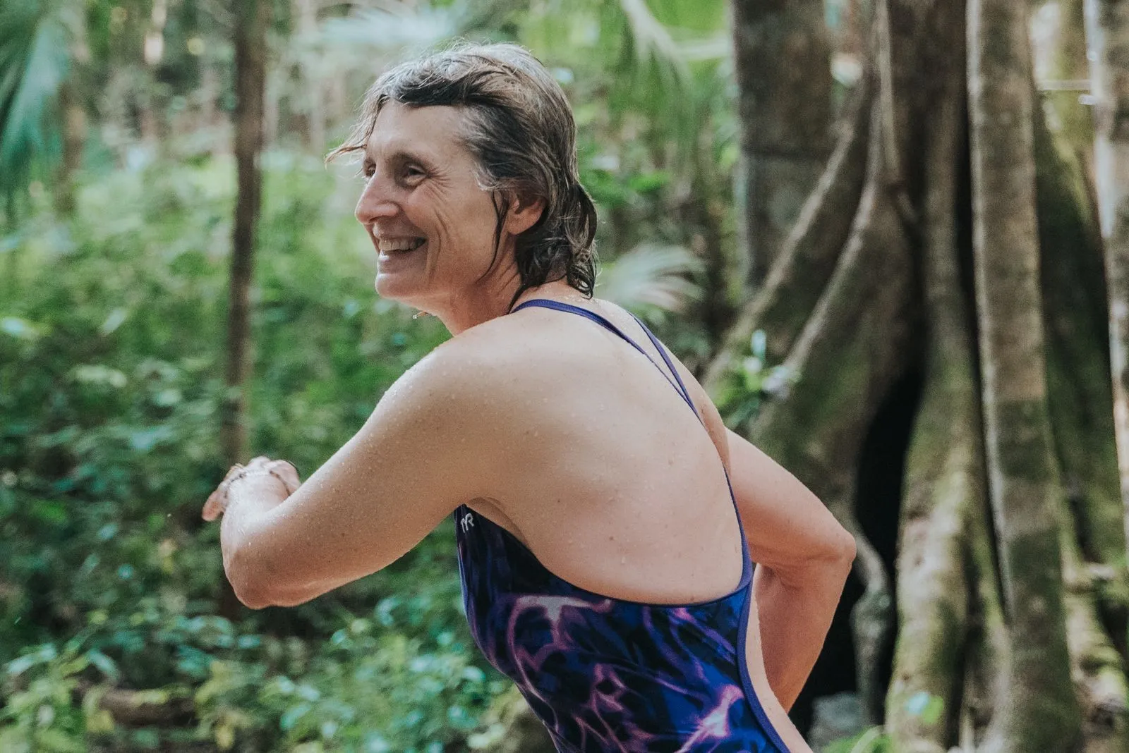 A smiling woman in a purple-patterned swimsuit turns toward the camera among dense jungle vegetation, Lunita Jungle Retreat