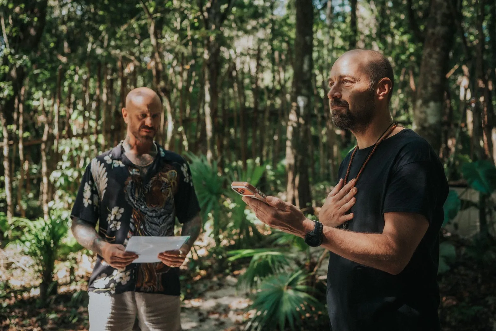 Two men stand on a jungle path dappled with overhead light, one in a patterned black shirt holding, Lunita Jungle Retreat