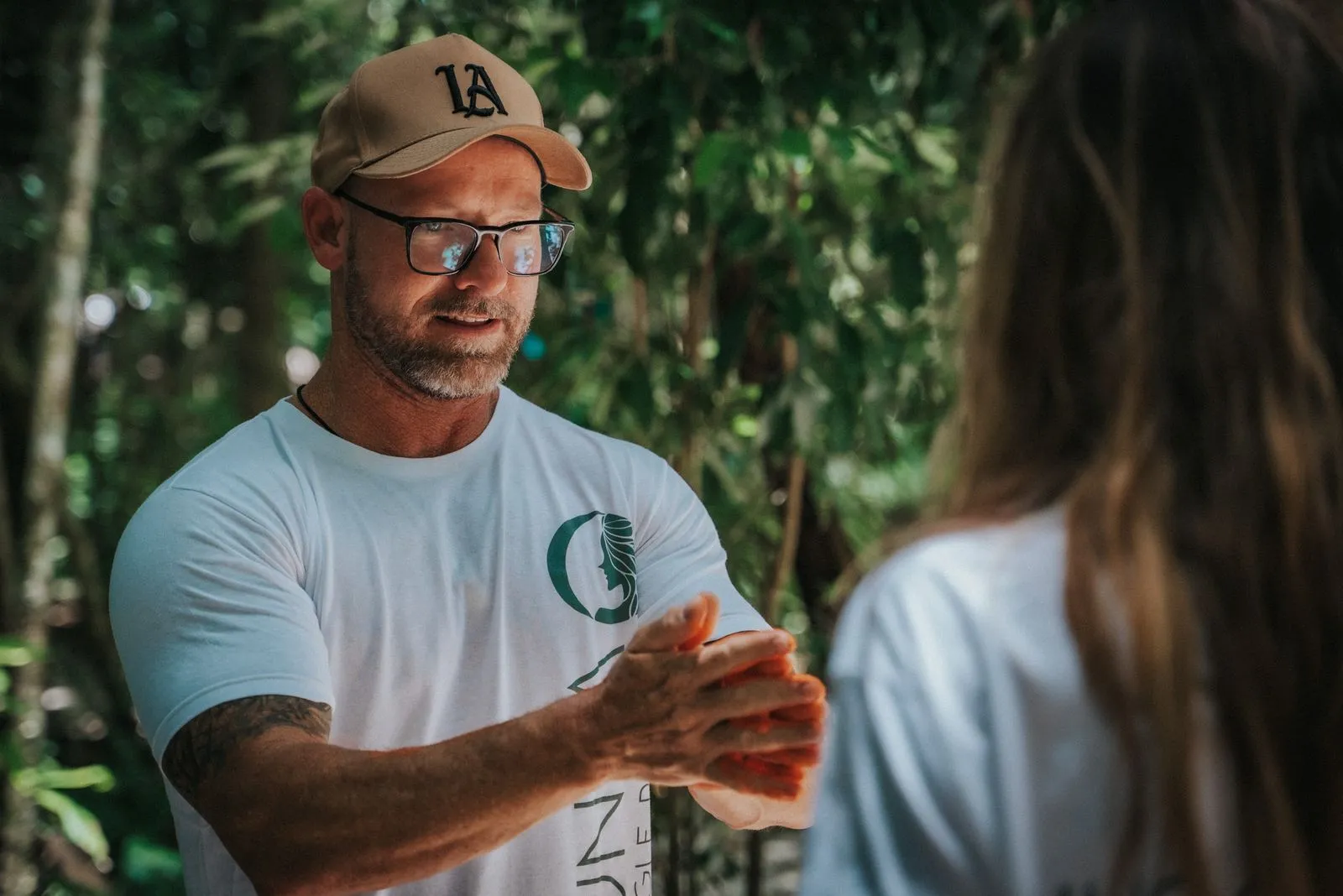 A man in a light blue t-shirt, tan cap, and sunglasses speaks gesturing with both hands to a woman, Lunita Jungle Retreat