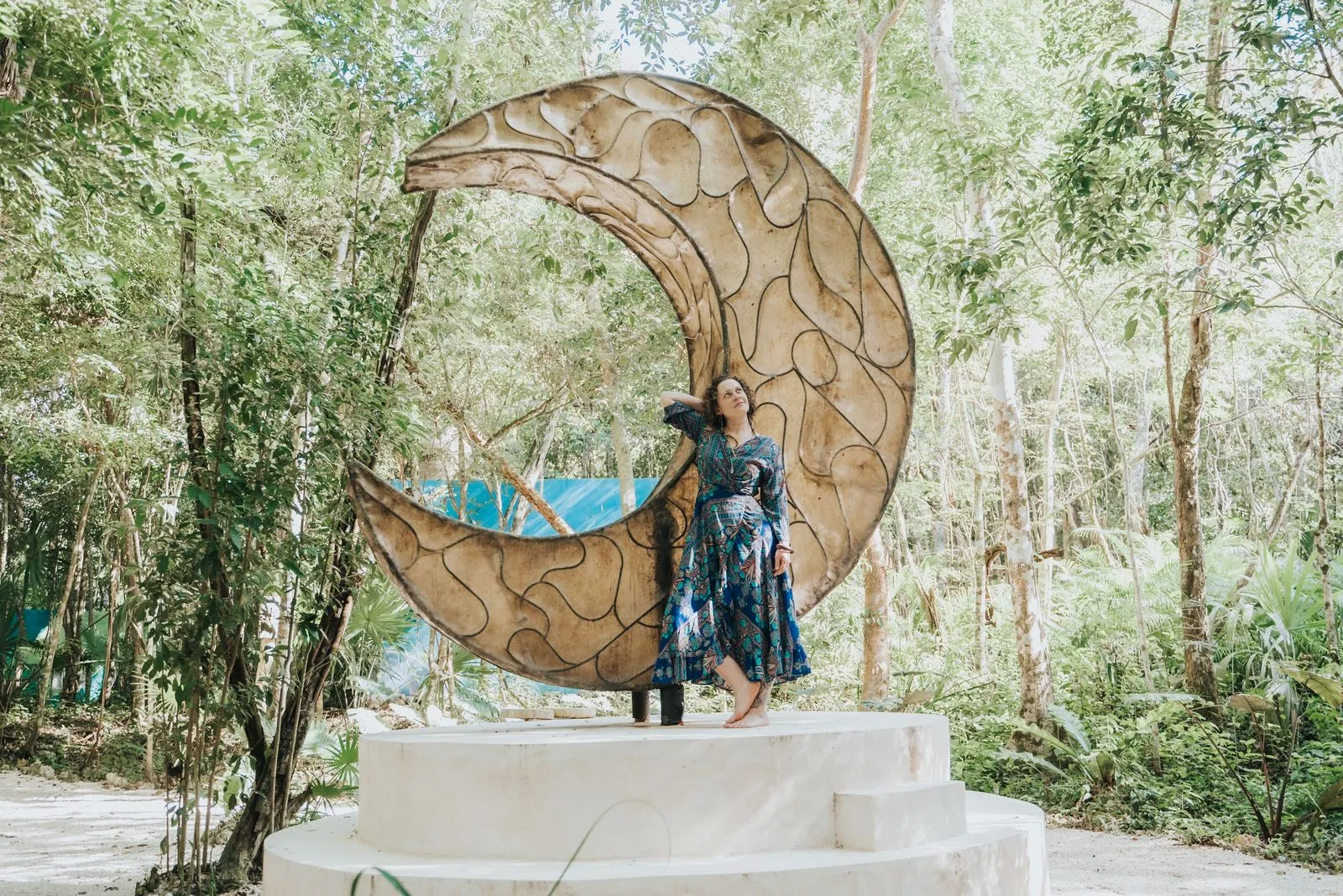 A woman in a navy floral dress stands on a white concrete platform beneath a large wooden spiral, Lunita Jungle Retreat