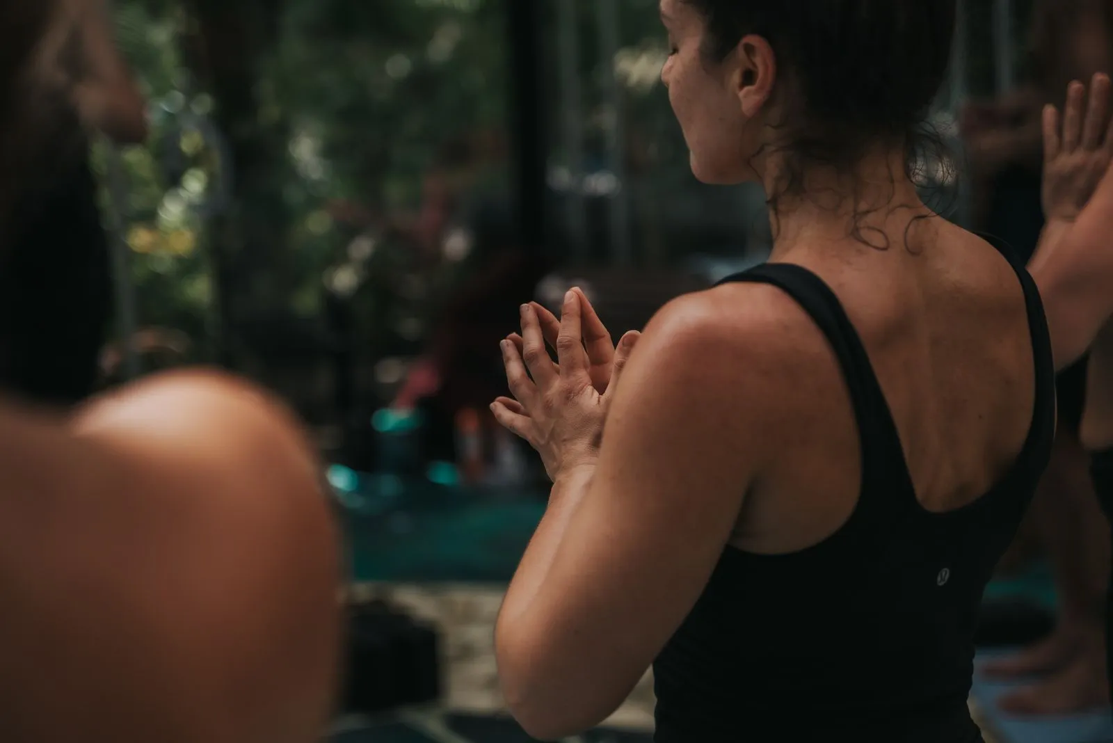 Woman in black tank top, hands at chest in prayer position, standing outdoors with dappled jungle, Lunita Jungle Retreat