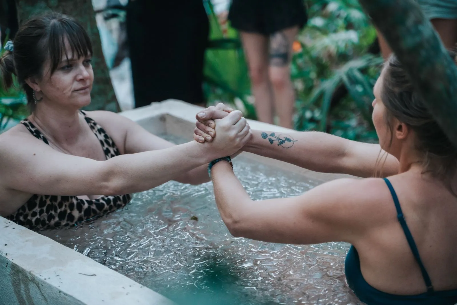 Two women grip forearms while submerged in a cenote pool, both wearing patterned swimwear, Lunita Jungle Retreat