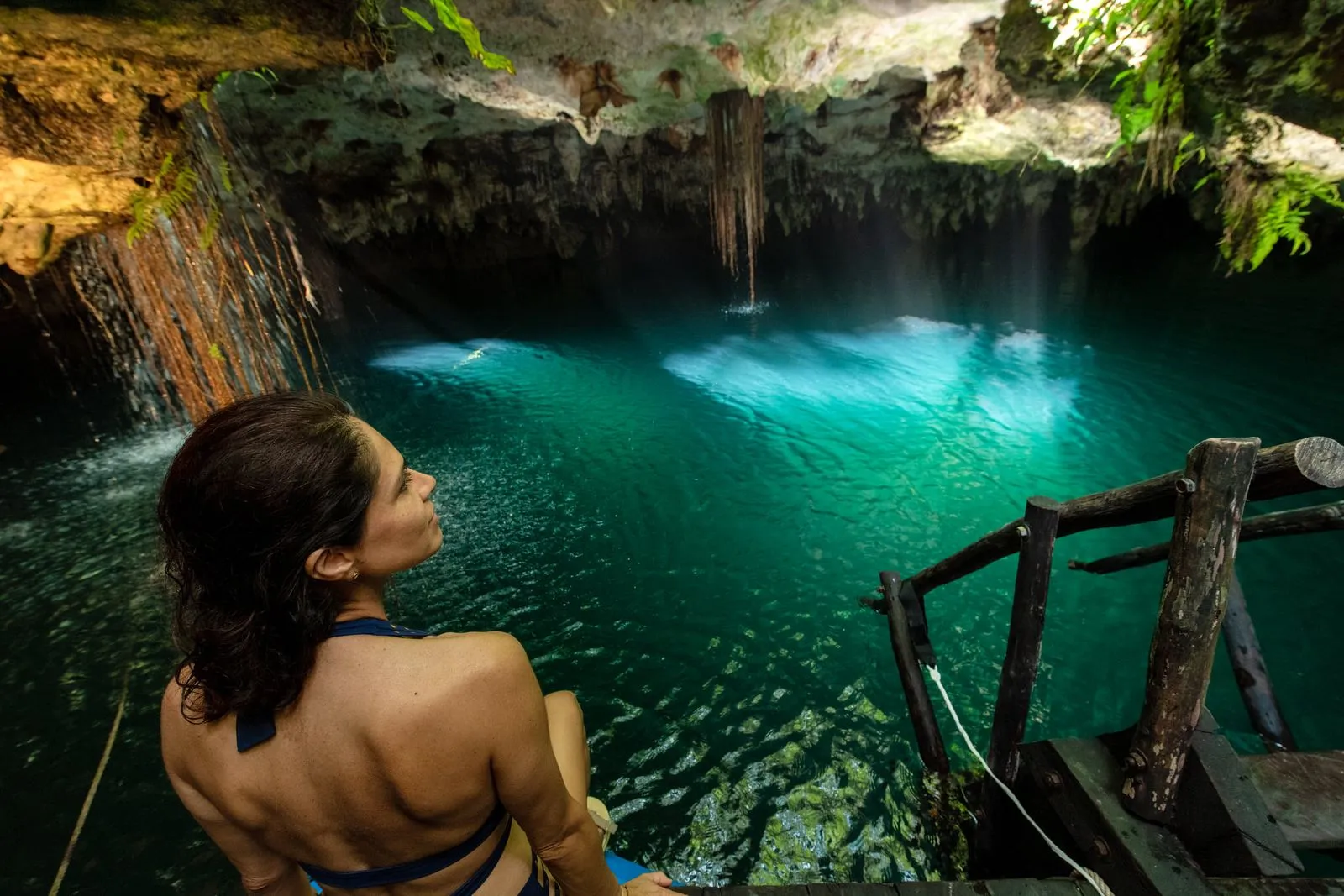 Woman in bikini stands on wooden platform gazing into turquoise cenote water, Lunita Jungle Retreat