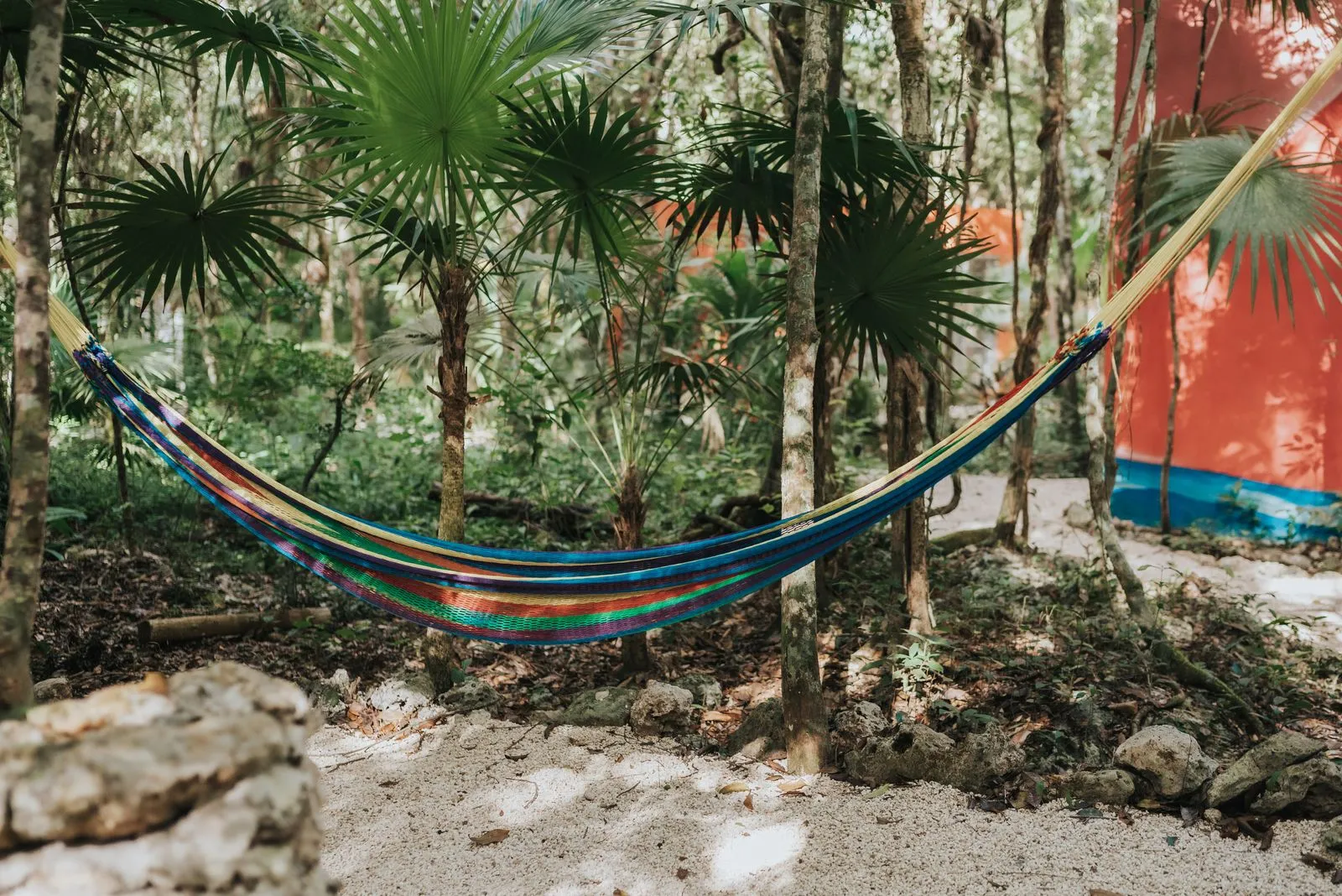 A woven hammock in blue, orange, and green stripes hangs between two thin jungle trees, Lunita Jungle Retreat