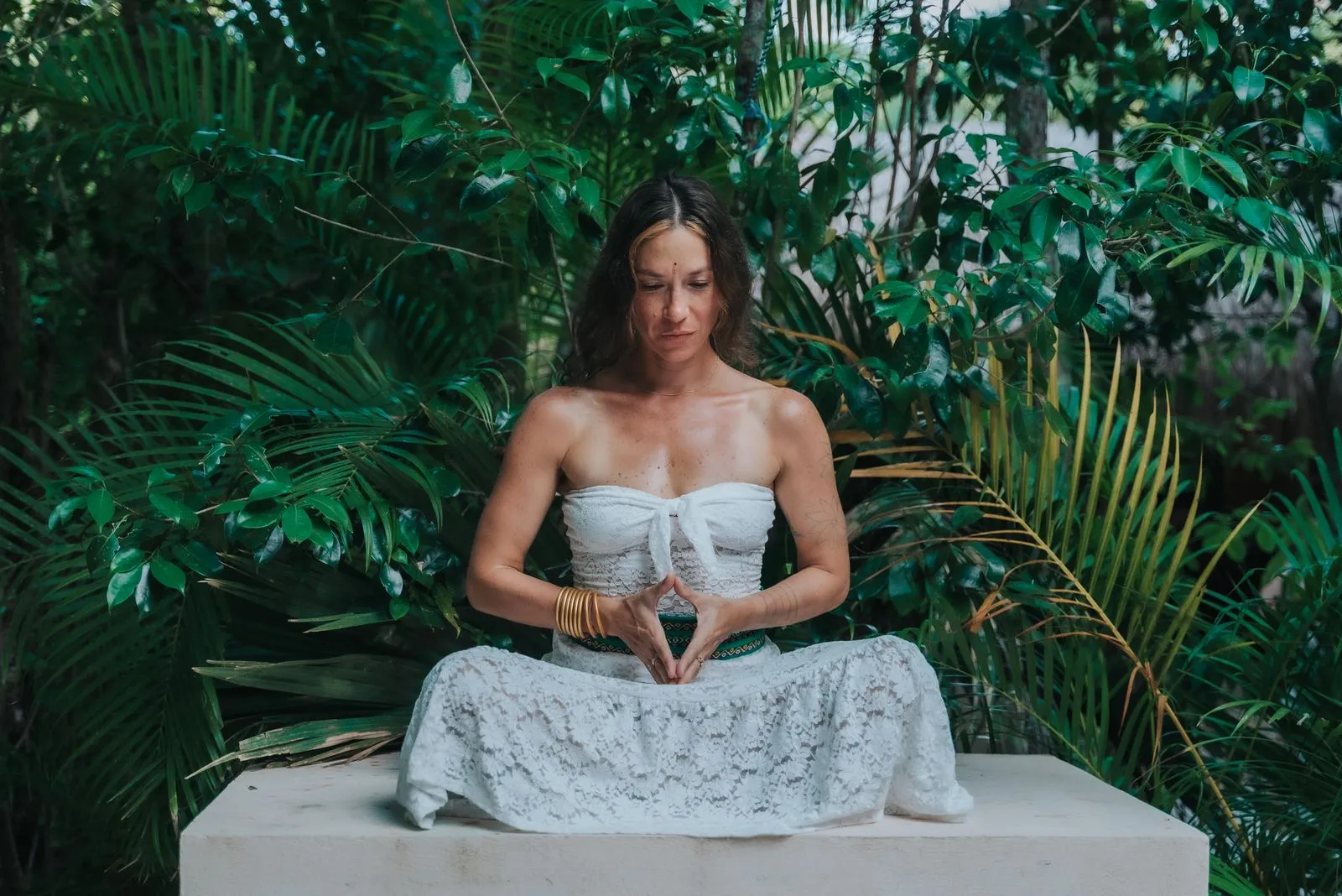 Woman in white lace dress seated on a concrete platform, hands folded at her center, Lunita Jungle Retreat