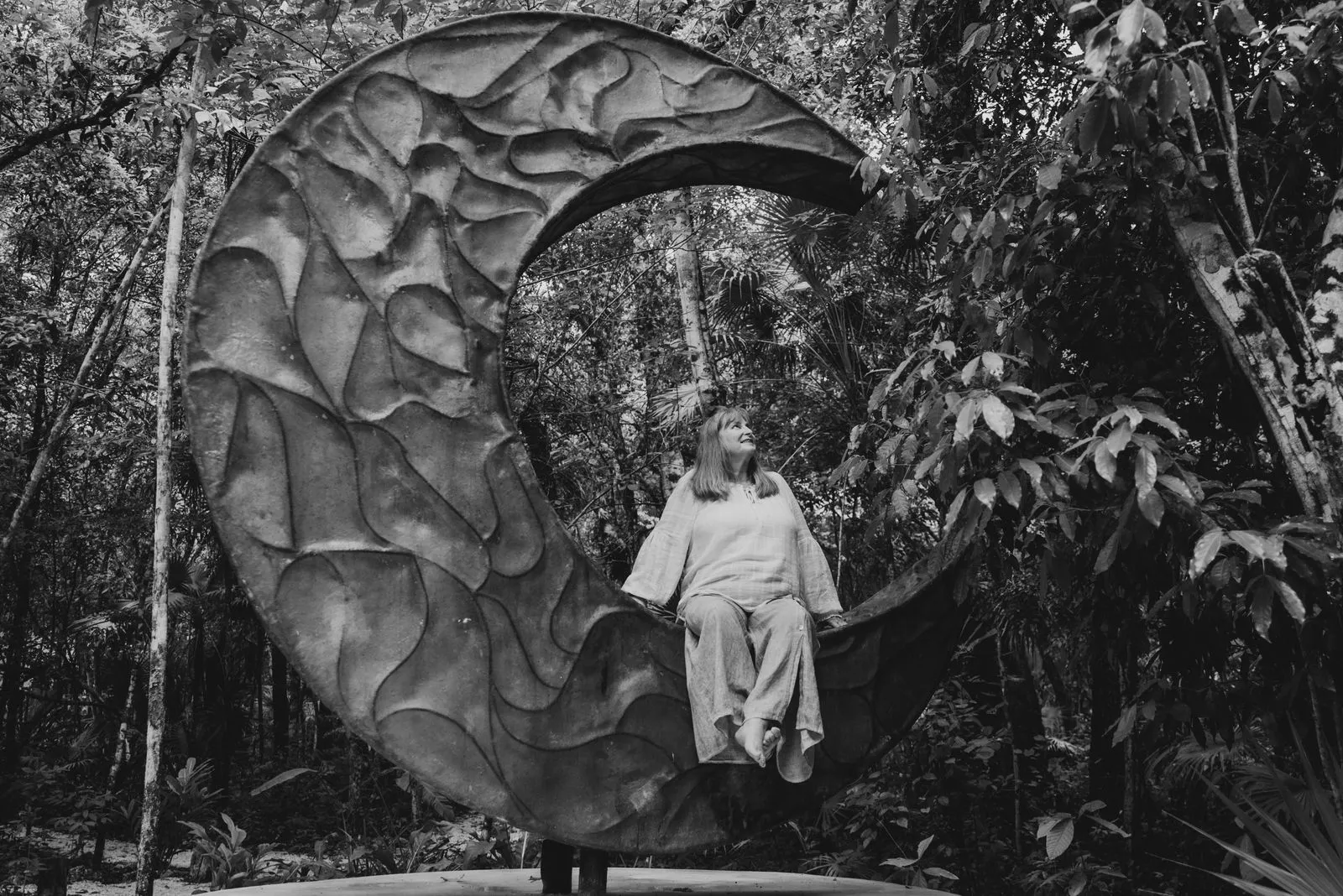 A woman in light linen clothing sits within a large spiral stone sculpture in dense jungle foliage, Lunita Jungle Retreat