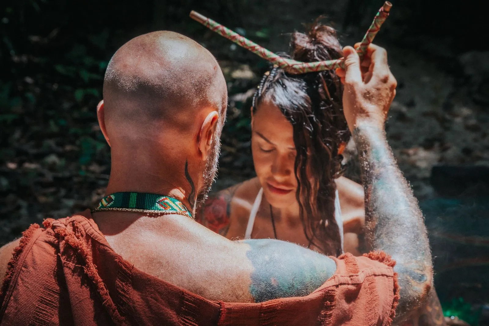 A man with shaved head and beaded collar, viewed from behind, helps a woman rinse her hair and body, Lunita Jungle Retreat