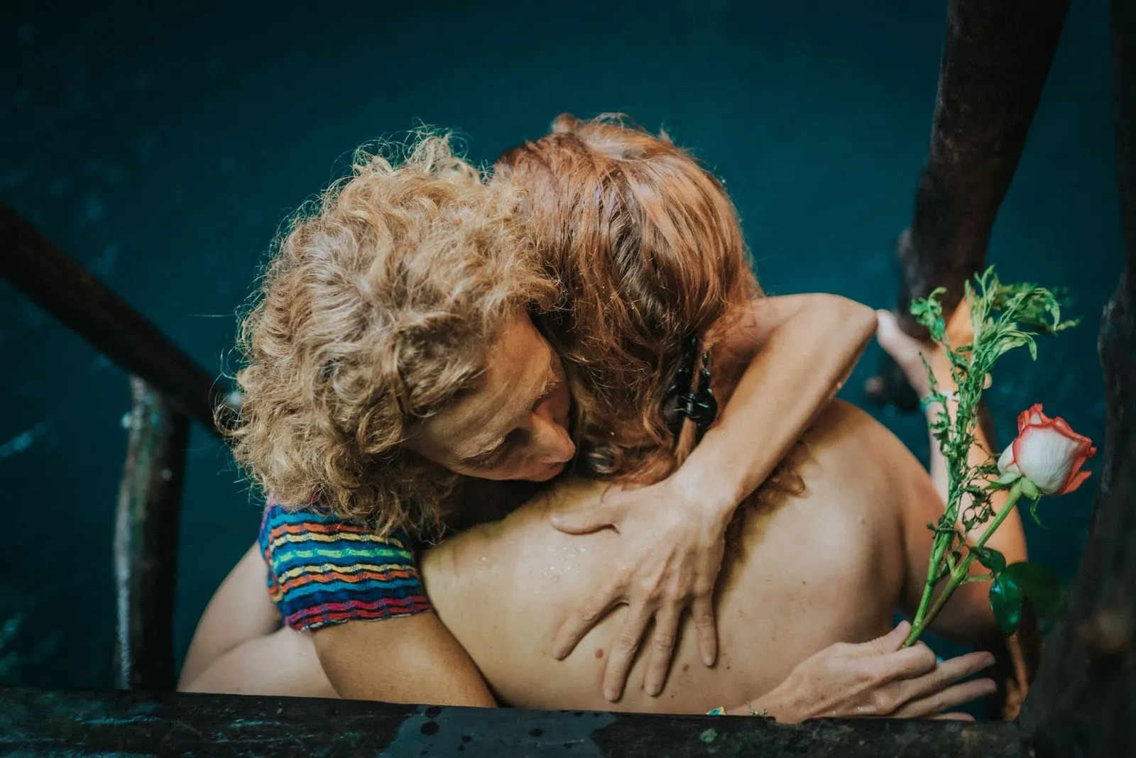 Two people embrace bare-chested against a deep teal wall, one wearing a woven rainbow-striped band, Lunita Jungle Retreat