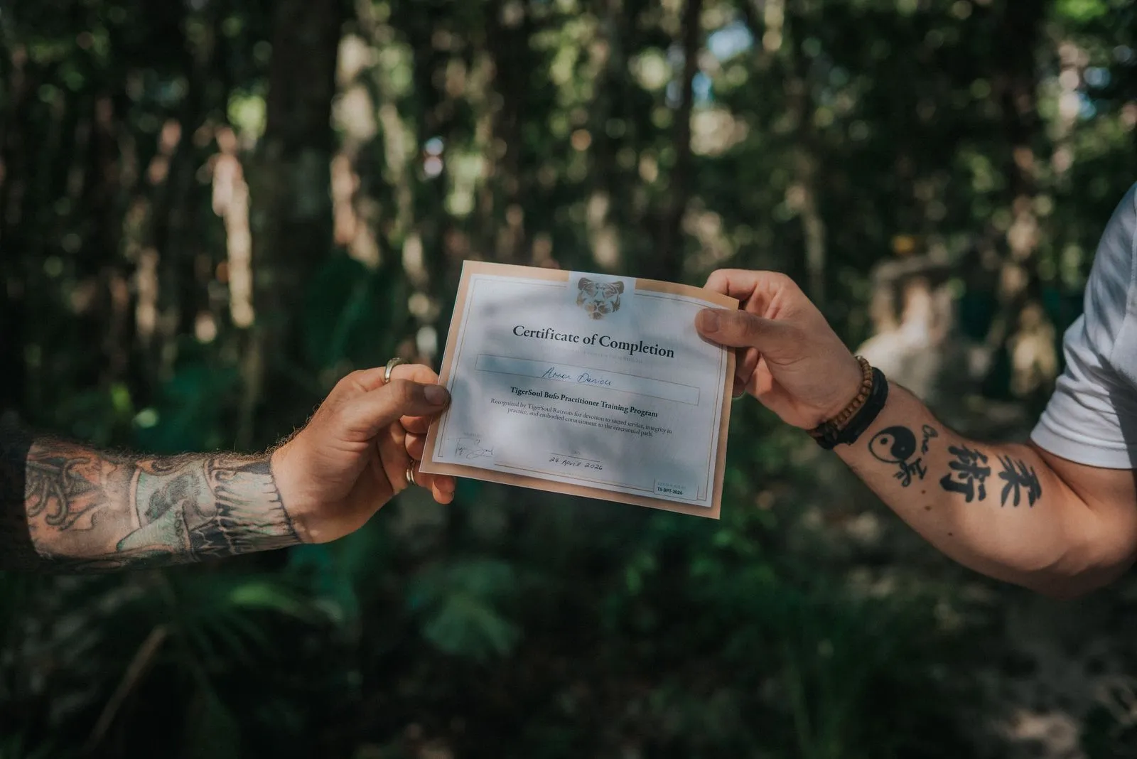 Two tattooed hands hold a Certificate of Completion between them in dappled jungle light, Lunita Jungle Retreat