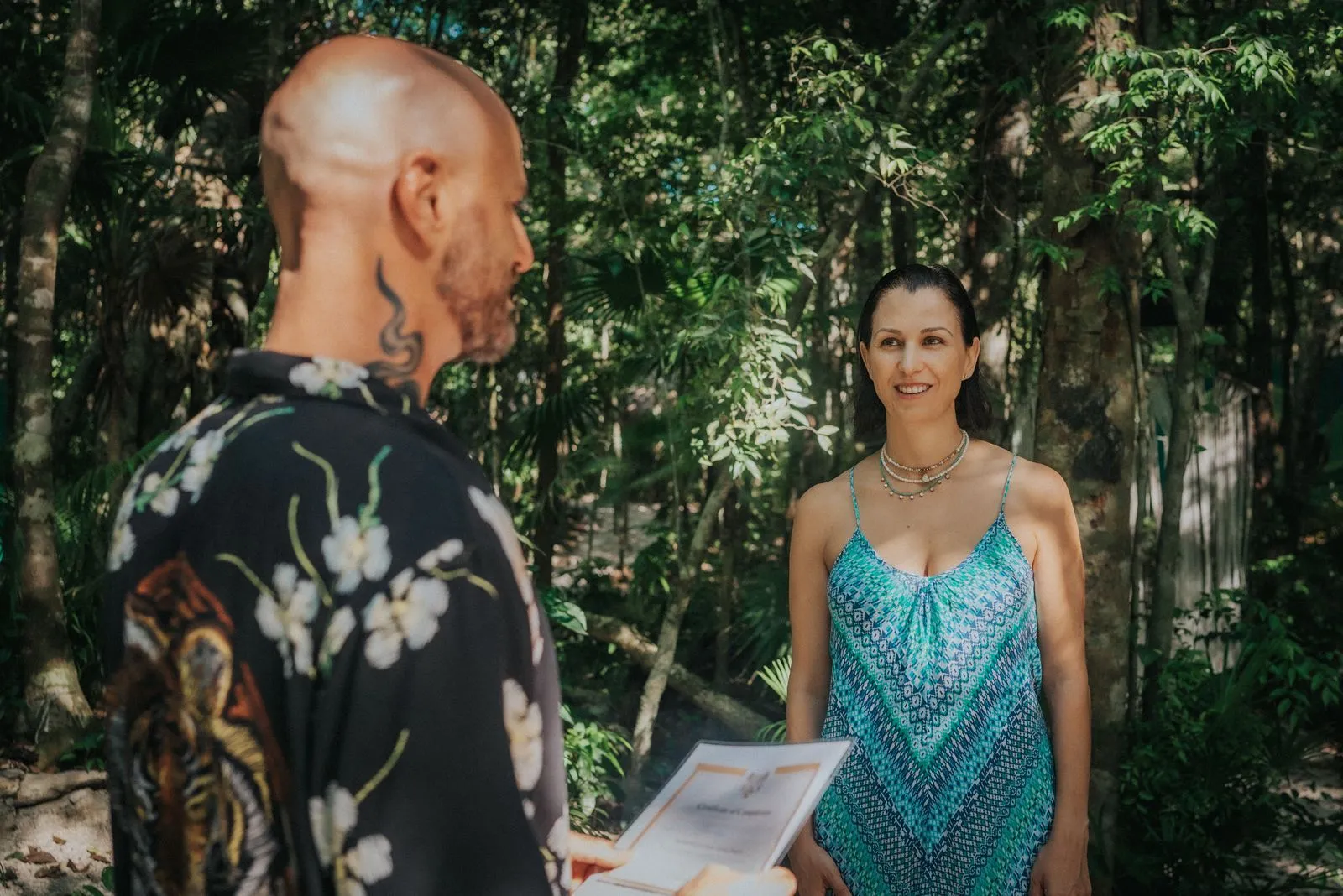 A man in a black floral shirt with a neck tattoo holds a clipboard facing a woman in a teal patterned, Lunita Jungle Retreat