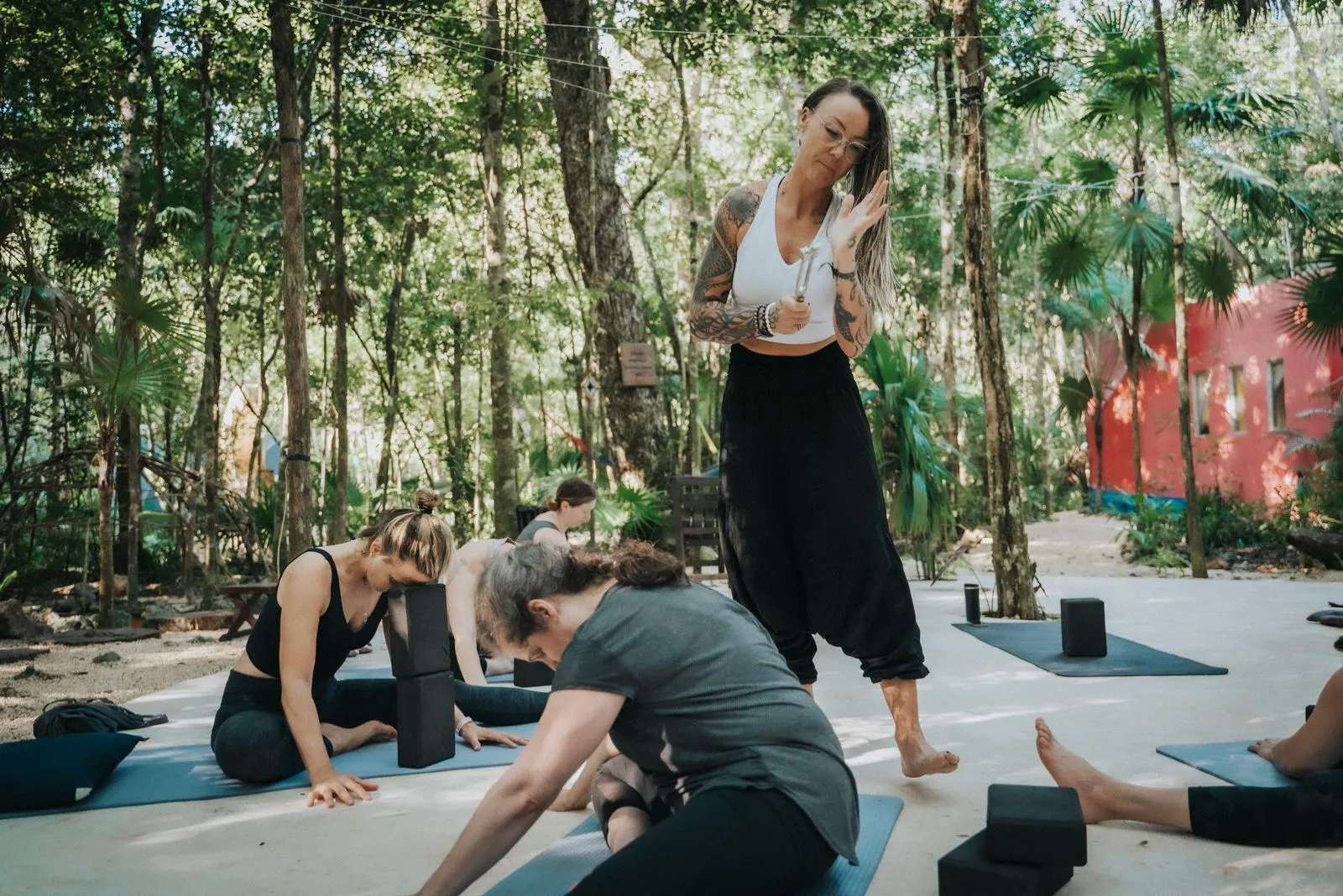 A yoga instructor in a white tank top and black pants guides three students on blue mats in a jungle, Lunita Jungle Retreat