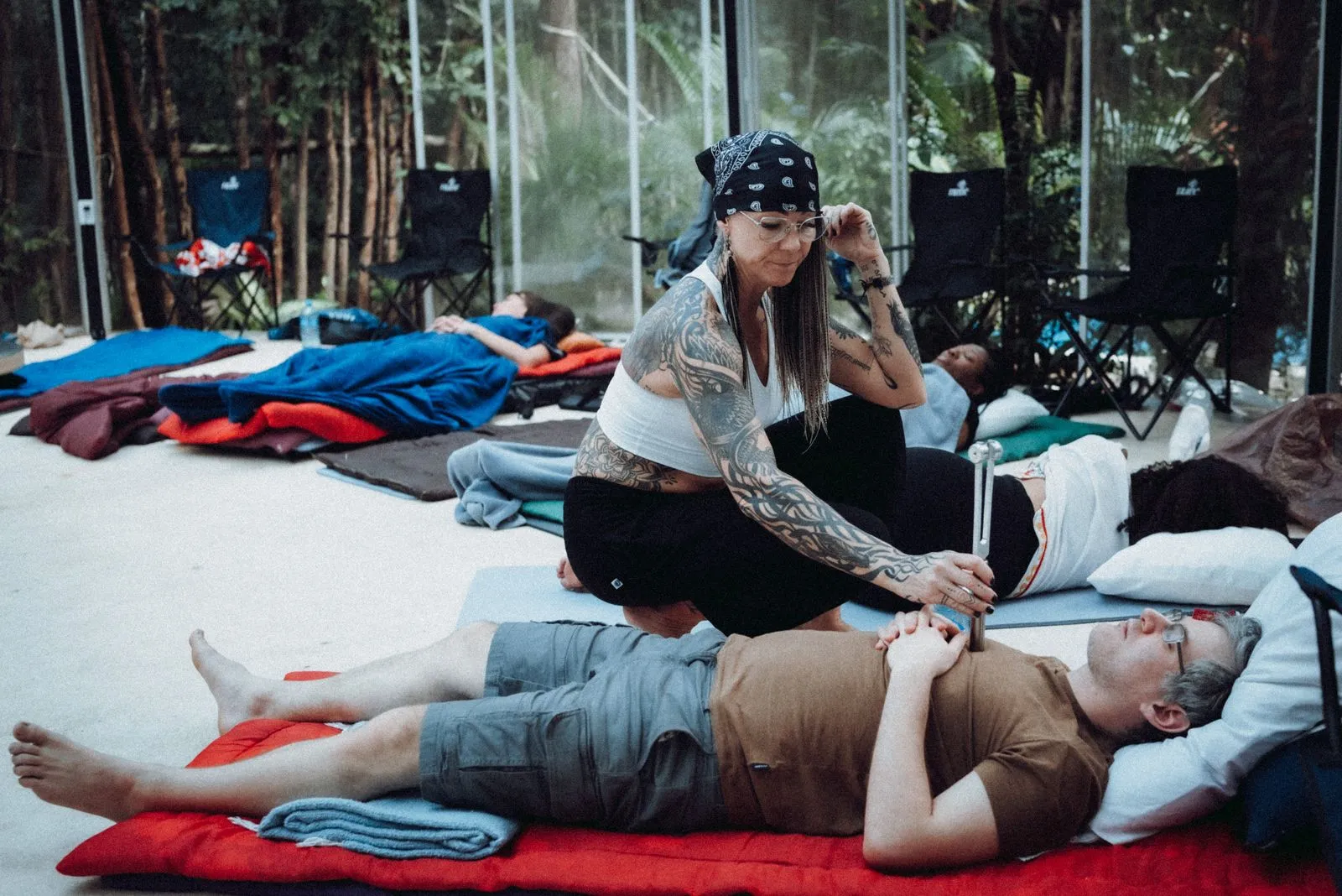 A tattooed facilitator in a white bandana and tank top adjusts a supine participant's arm during, Lunita Jungle Retreat