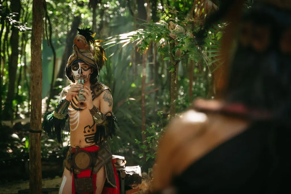 A performer in skull face paint, feathered headdress, and body tattoos holds a wooden flute in a dense, Lunita Jungle Retreat