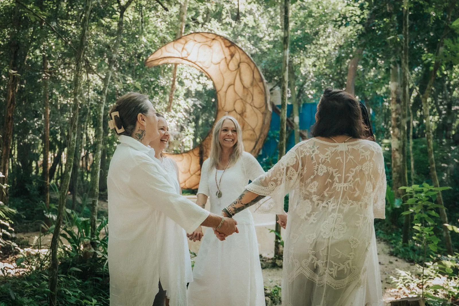 Three women in white linen stand hand-to-hand on a jungle path beneath a large wooden crescent moon, Lunita Jungle Retreat