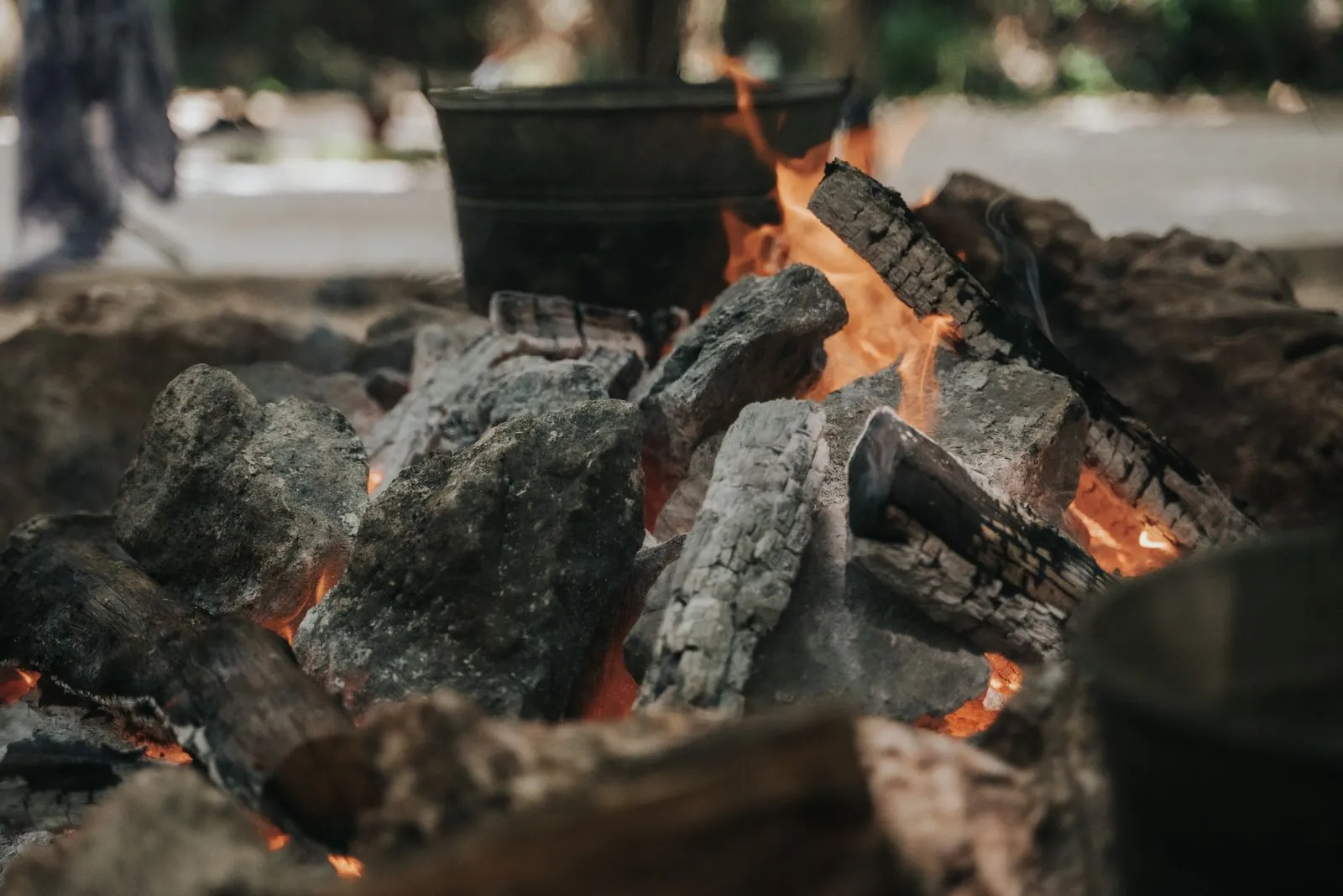 Close-up of a live fire pit with charred logs and glowing orange flames, a black metal pot visible in soft focus behind. People are blurred in the background on a daytime path.