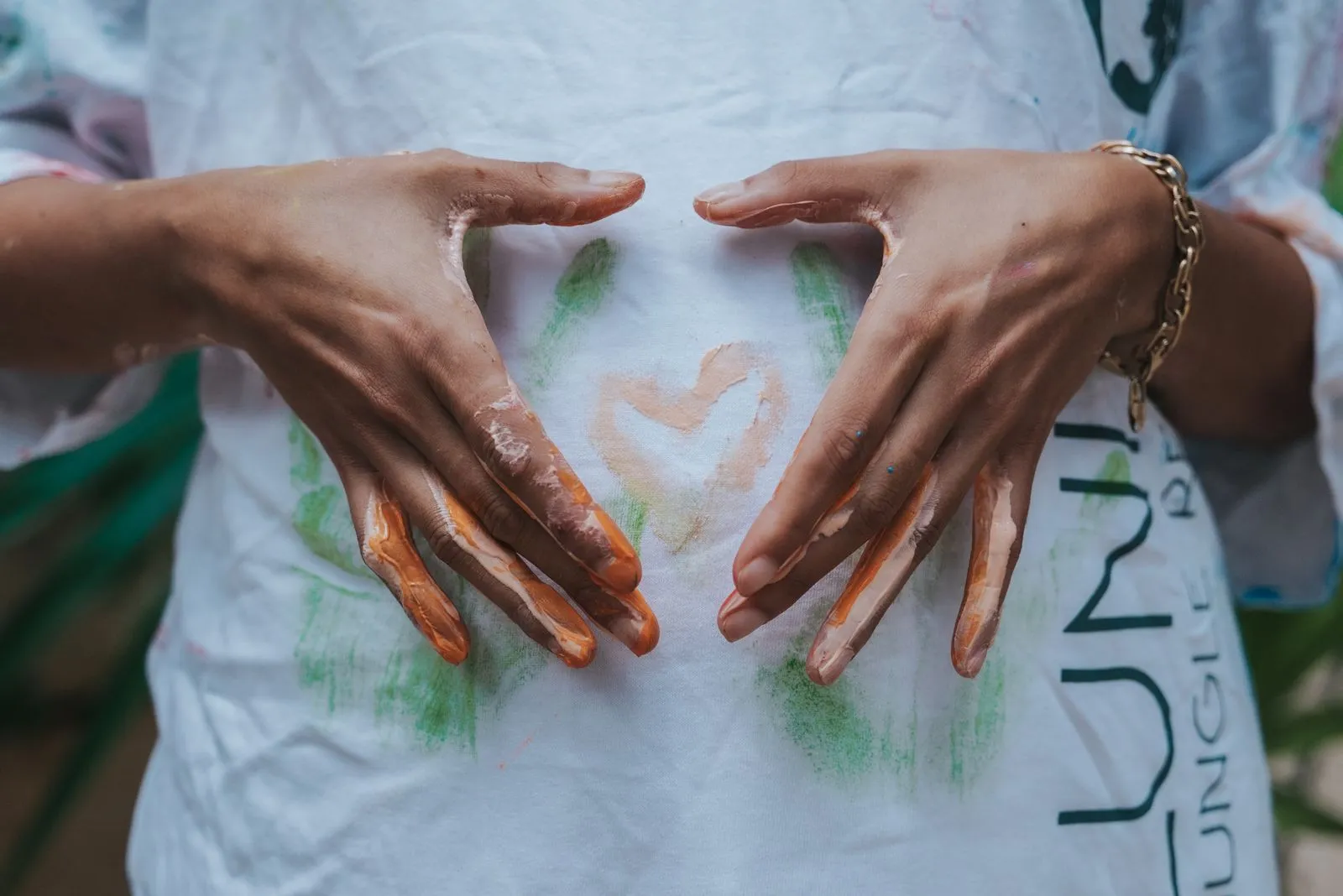 Two hands framing a heart shape on a white t-shirt stained with cacao and green plant matter. A gold, Lunita Jungle Retreat
