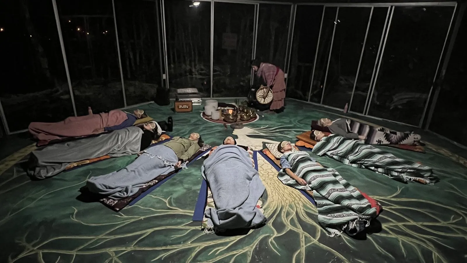 Eight people lie on sleeping bags across a green patterned floor in a glass-walled palapa at night, Lunita Jungle Retreat