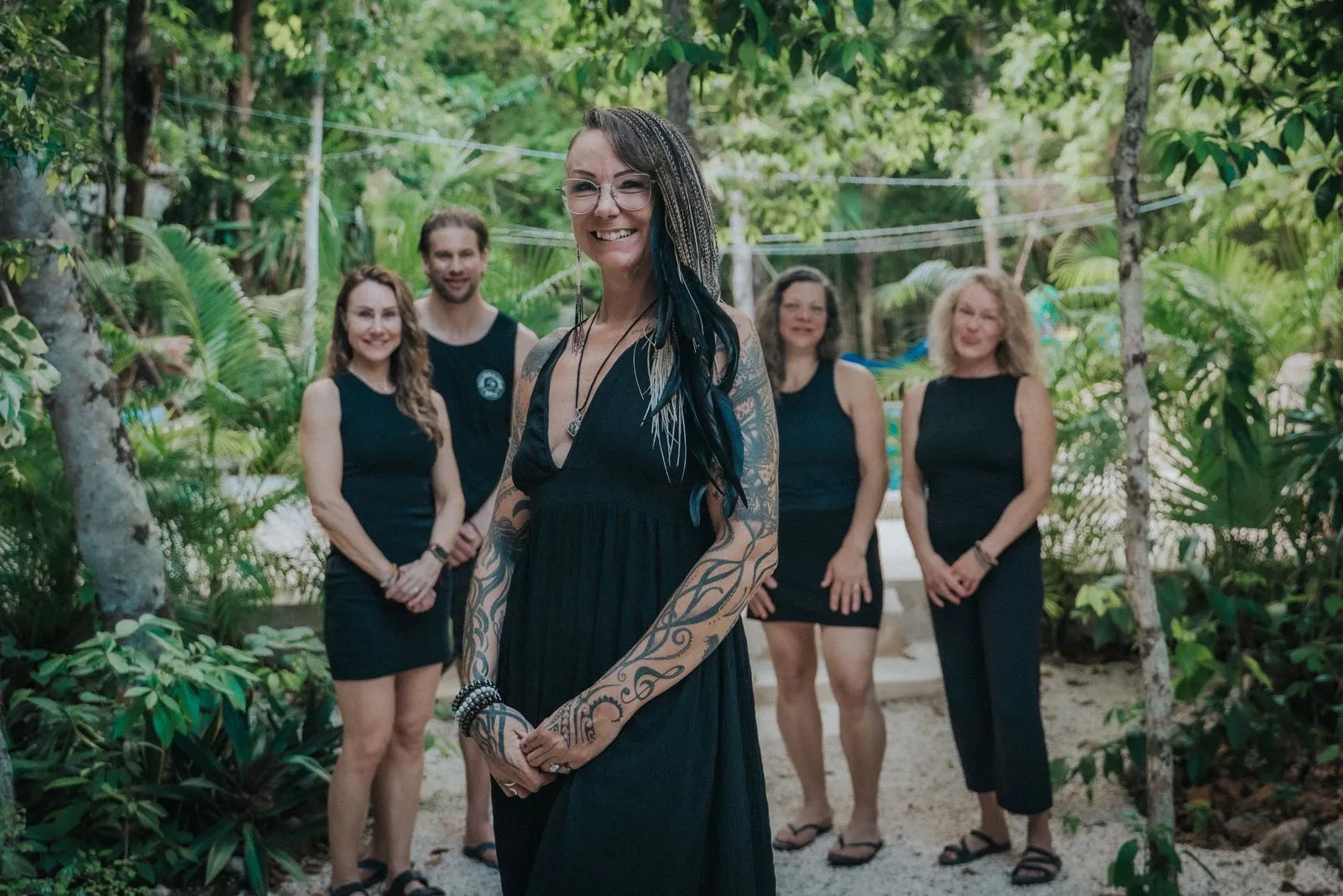 A woman with black braids, glasses, and full-sleeve tattoos stands in the foreground wearing a black, Lunita Jungle Retreat