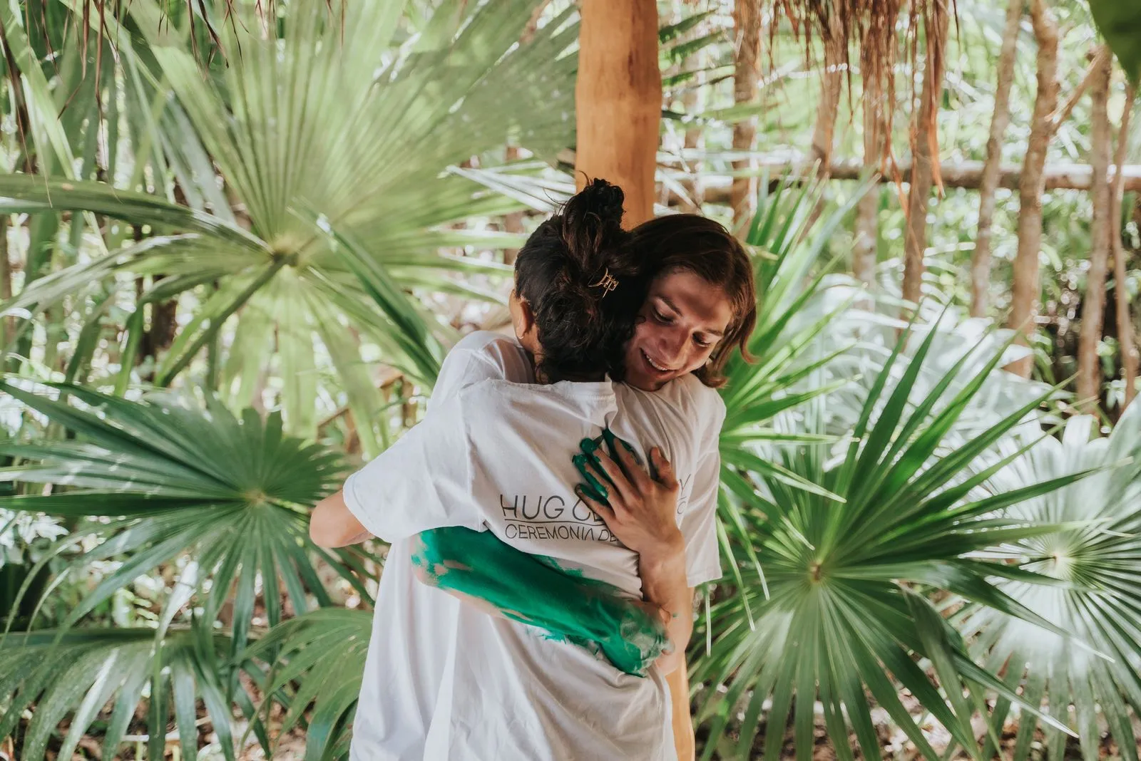 Two people embrace in dappled jungle light, surrounded by tall palms and dry fronds. The person, Lunita Jungle Retreat