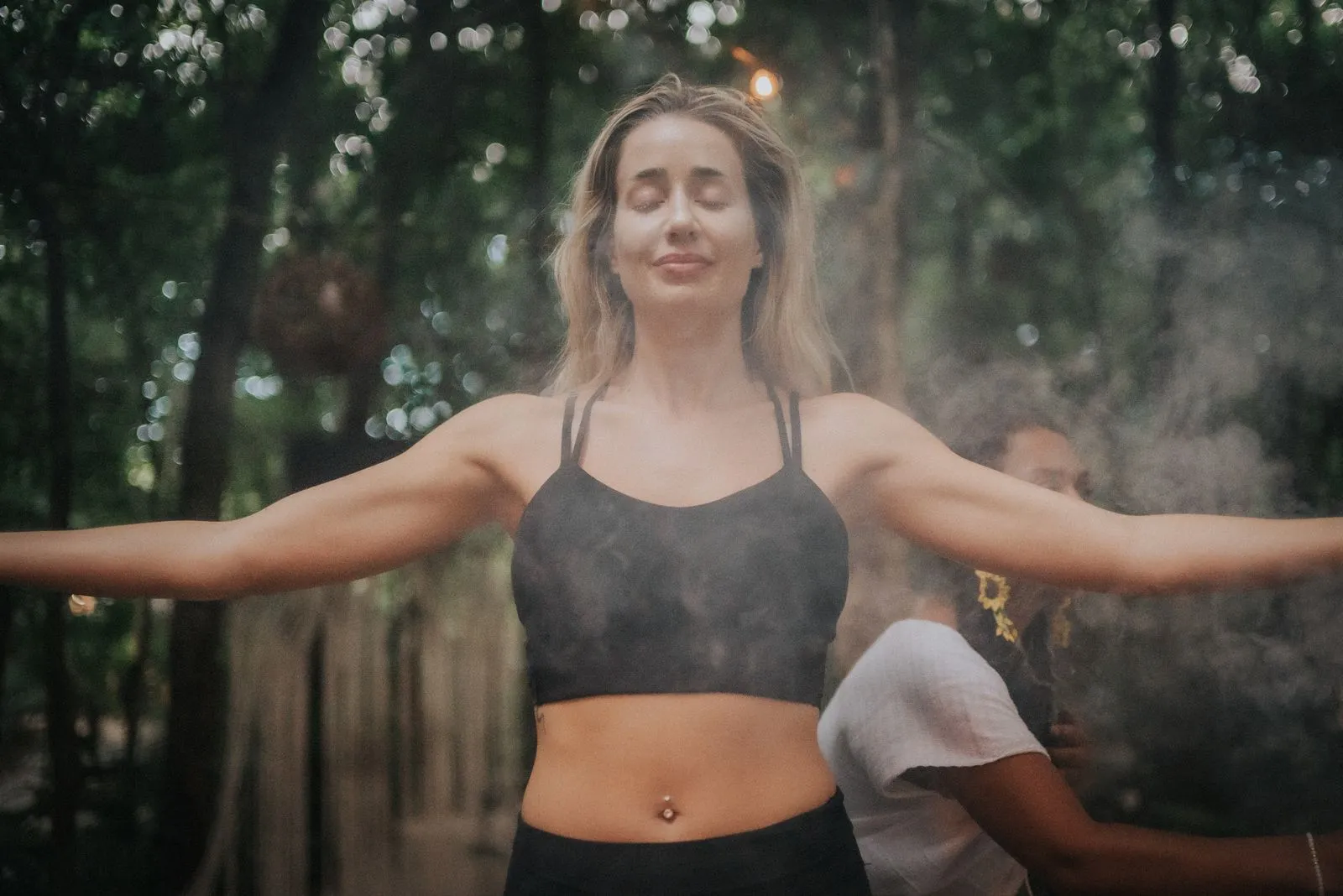 A woman in a dark sports bra stands with arms extended in an open chest pose on a wooden platform, Lunita Jungle Retreat
