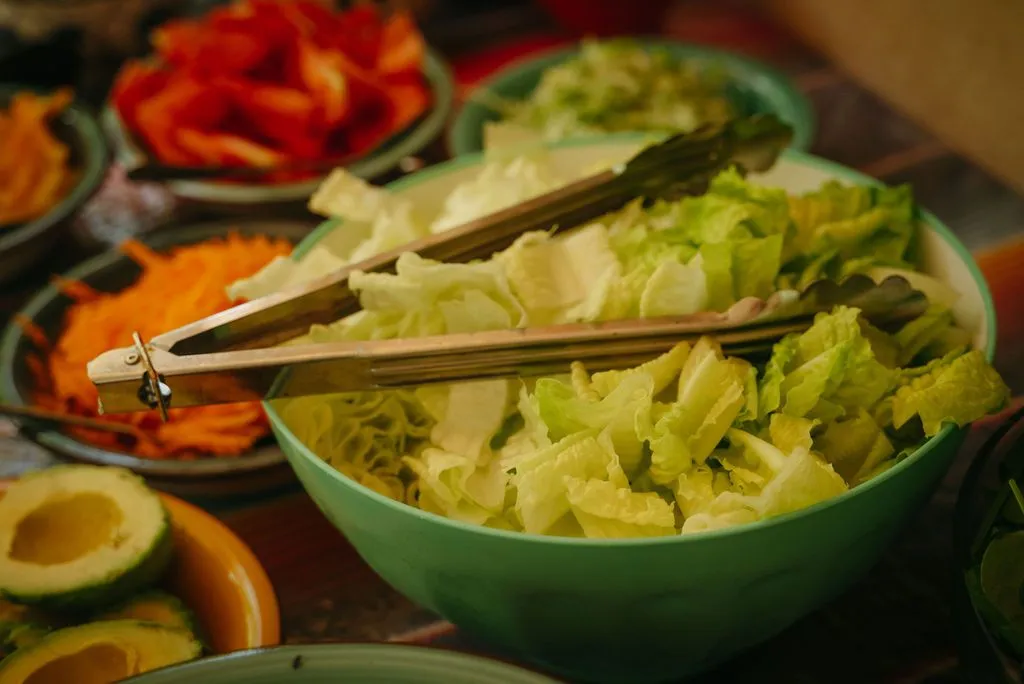 A bright-green ceramic bowl of fresh lettuce with wooden serving tongs rests on a wooden table, Lunita Jungle Retreat