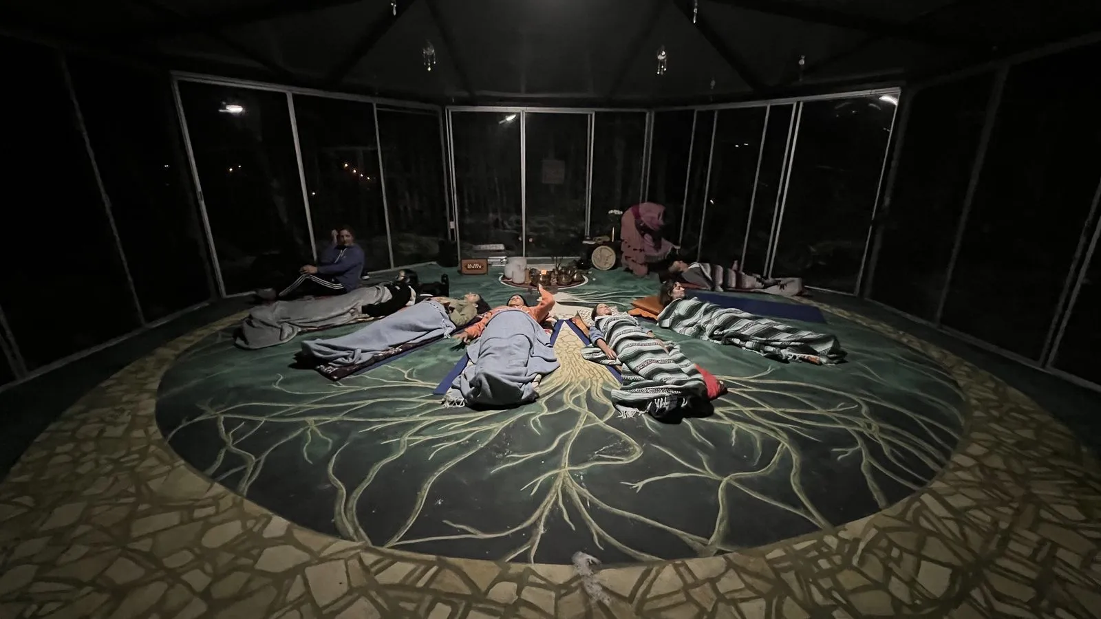 Six people lie and recline on a large green fabric printed with root-like patterns across the floor, Lunita Jungle Retreat