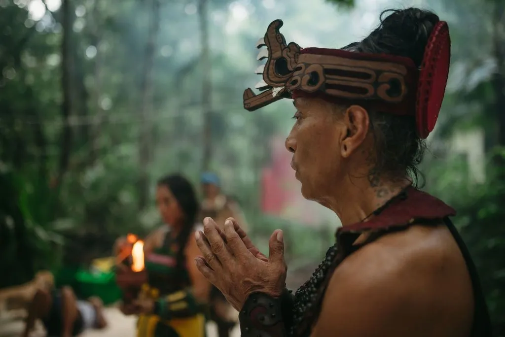 An elder in profile wears an ornate wooden headdress with a carved serpent motif and red fabric, Lunita Jungle Retreat