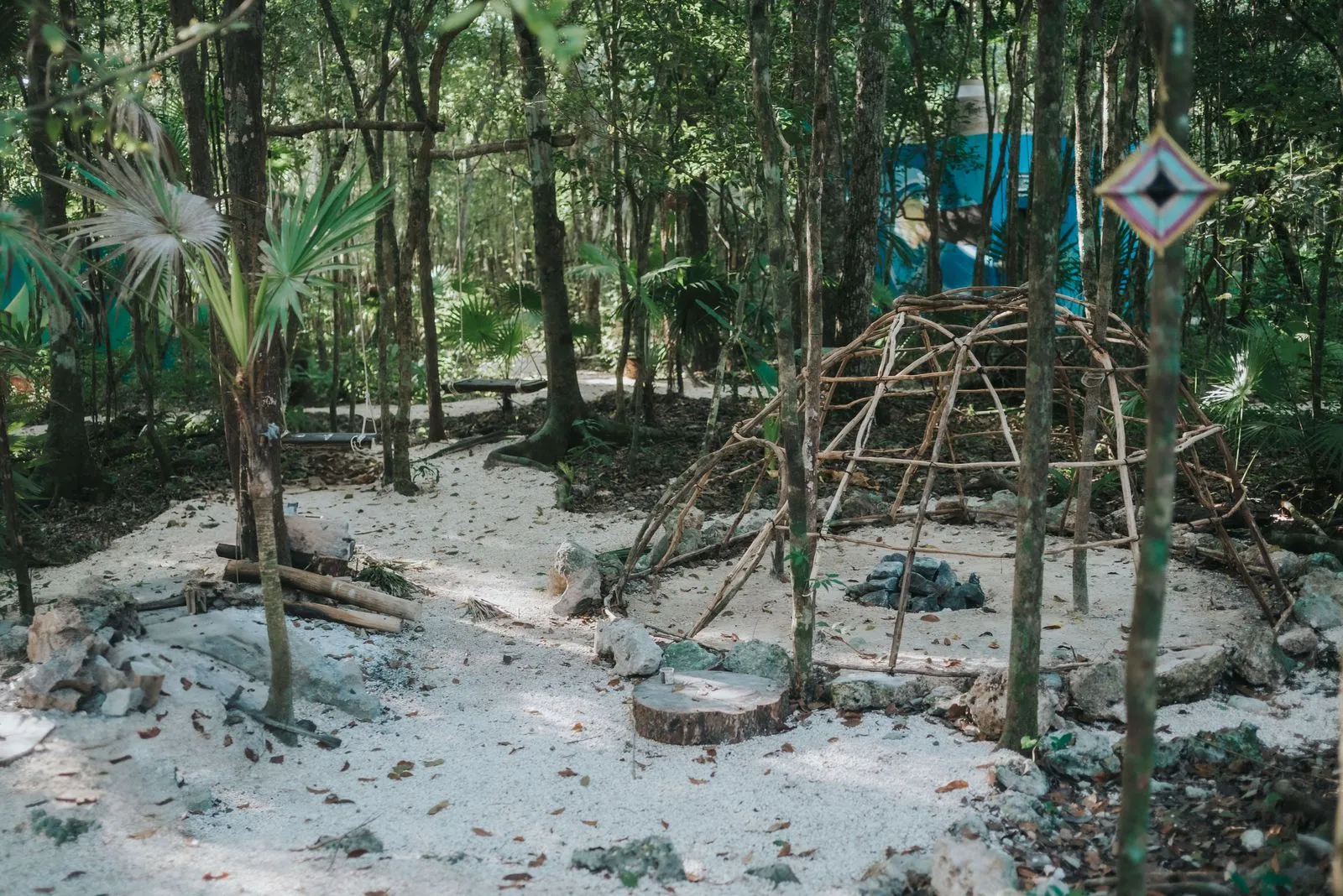 Sandy jungle clearing with woven stick structures and a blue fabric shelter visible through dense, Lunita Jungle Retreat