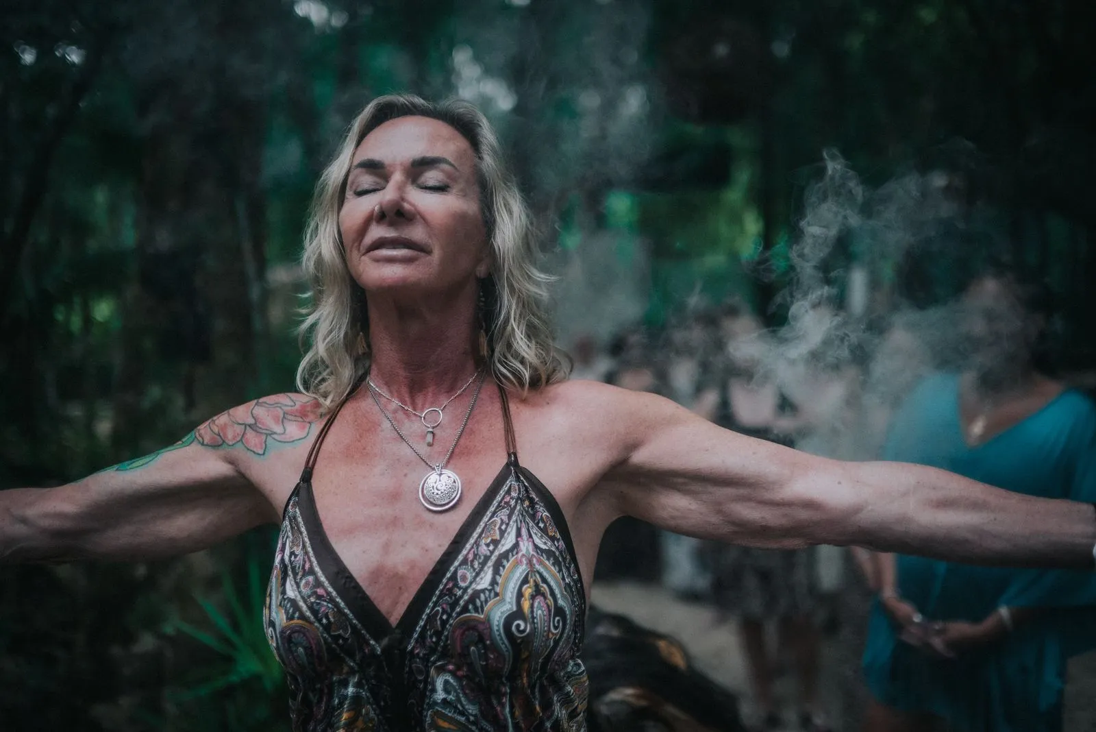 A woman with long blonde hair and a shoulder tattoo stands with arms spread wide in the jungle, Lunita Jungle Retreat