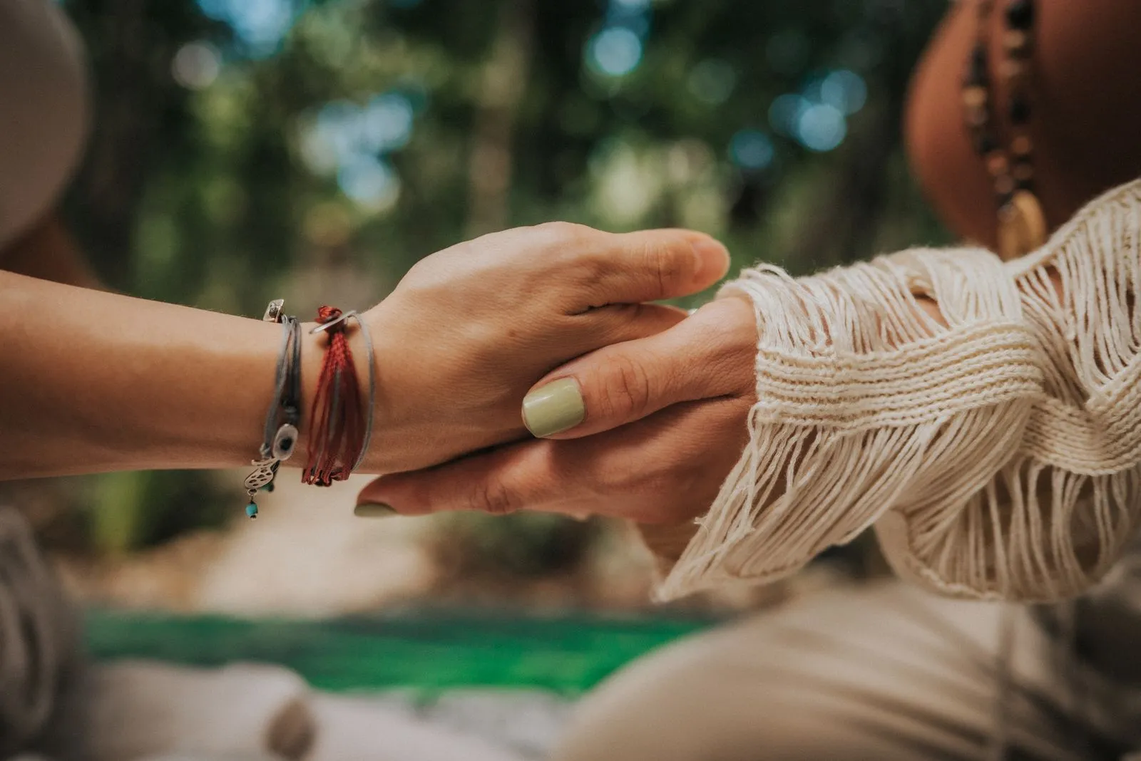 Two hands meet in a gentle clasp outdoors, one wearing a cream cable-knit sleeve and beaded bracelets, Lunita Jungle Retreat