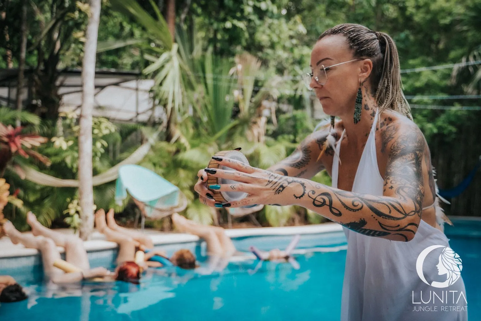 A facilitator in a white halter top and beaded earrings stands at the pool's edge, Lunita Jungle Retreat