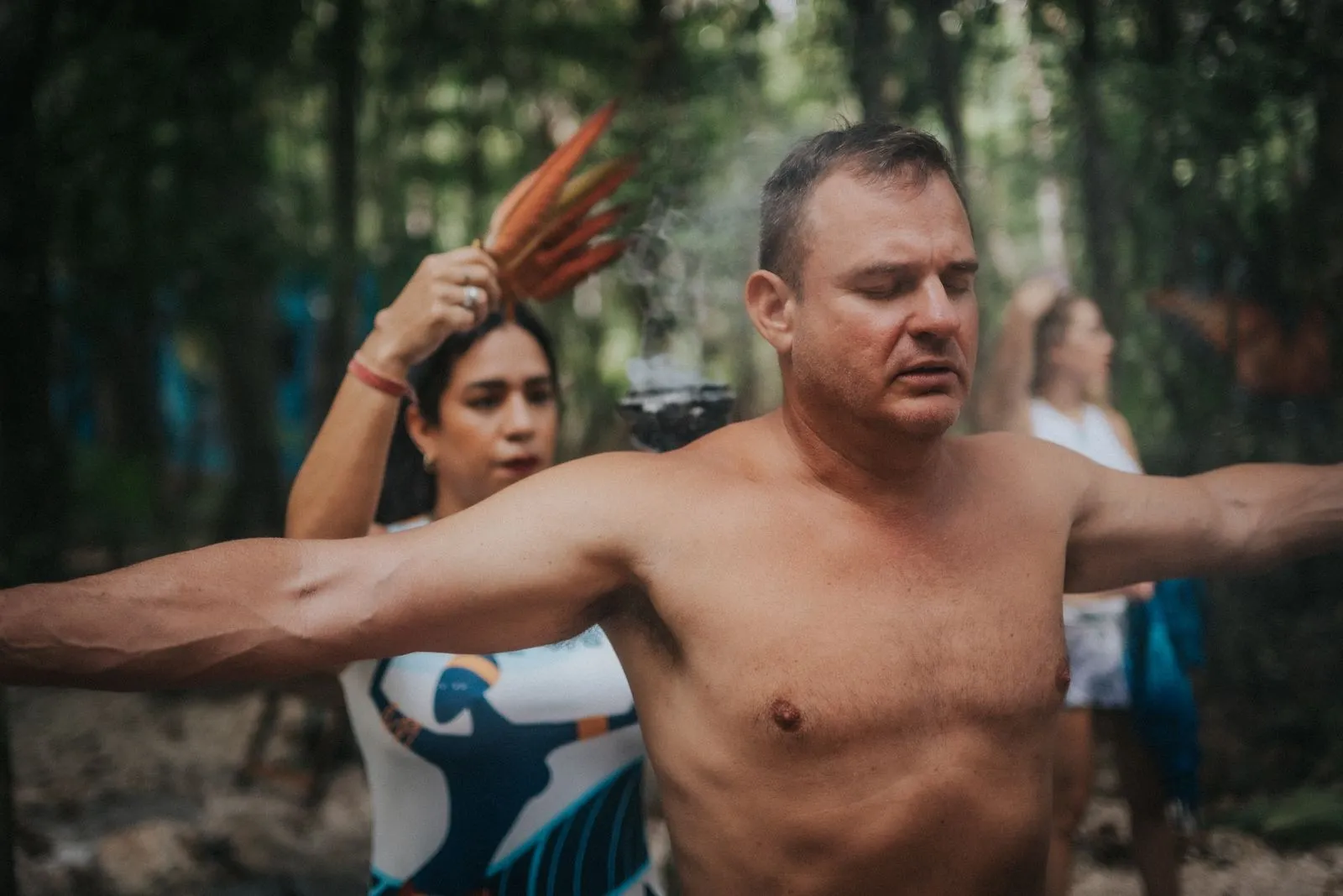 Shirtless man with arms extended holds red feathers while standing in dappled jungle light, Lunita Jungle Retreat