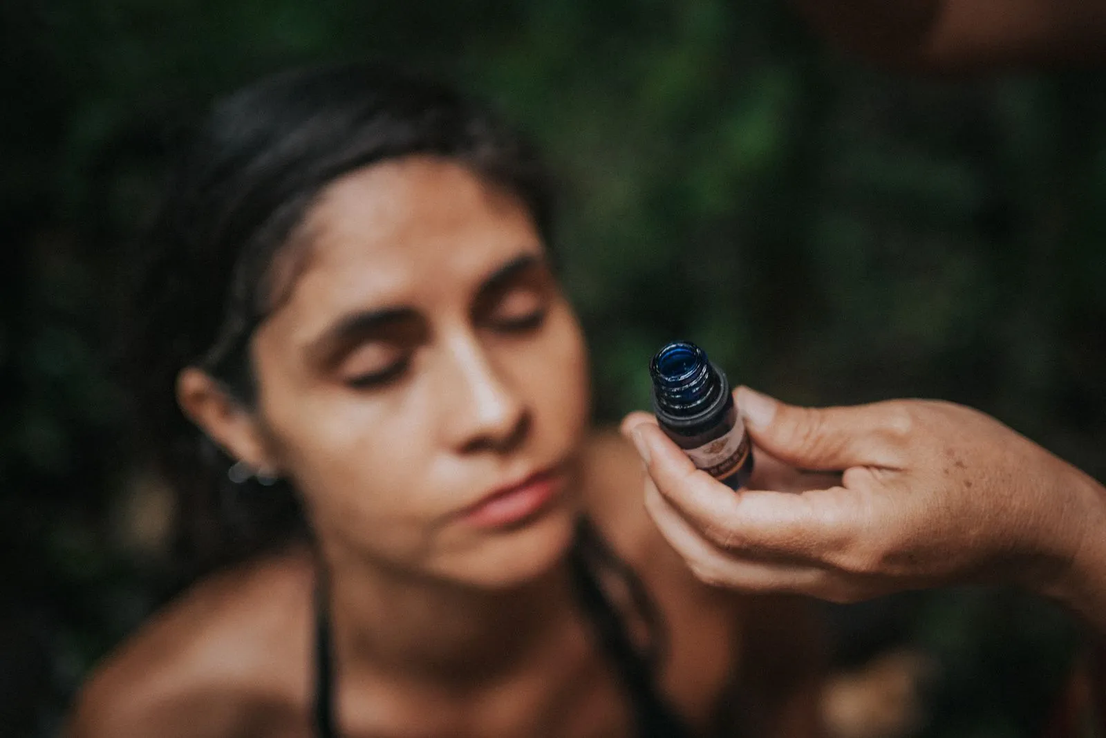 A woman with eyes closed receives a small dark glass bottle held by a hand in the foreground, Lunita Jungle Retreat