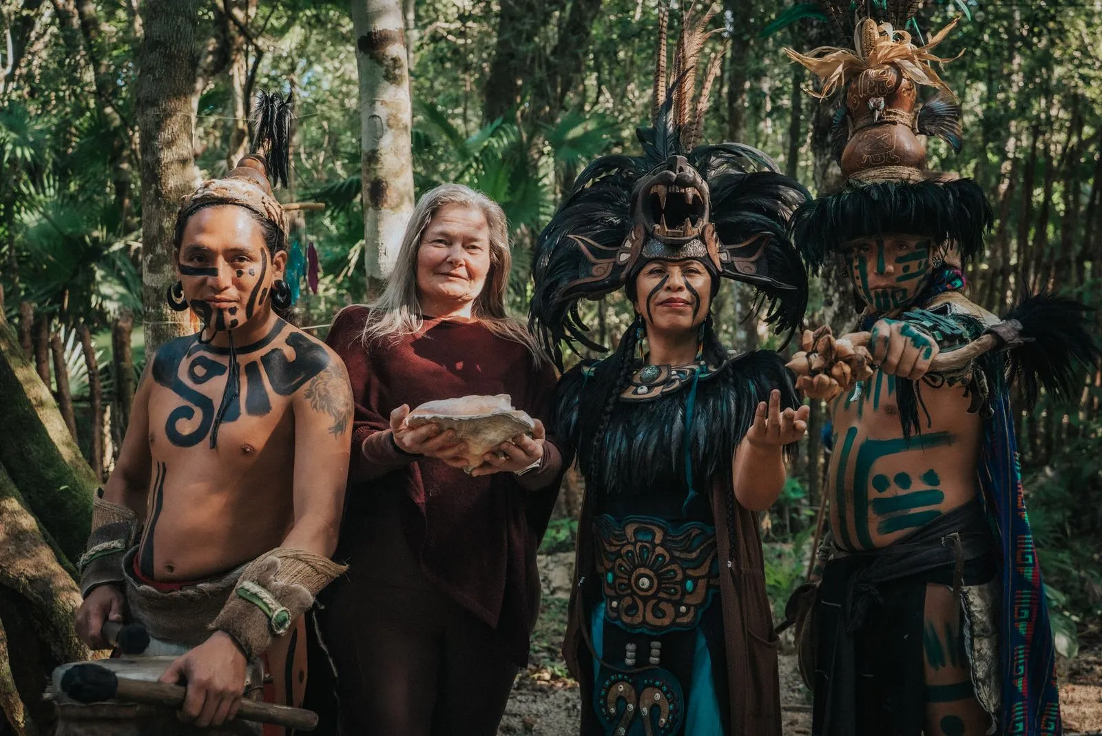 Four people in ceremonial dress and body paint stand in a jungle clearing with tall palms and trees, Lunita Jungle Retreat
