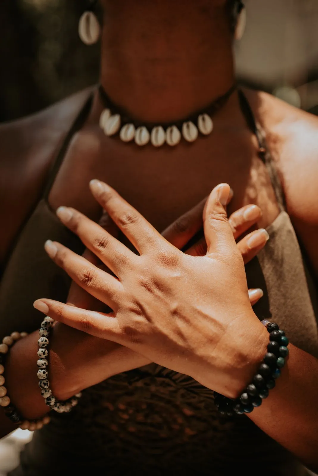 Extreme close-up of two brown-skinned hands crossed over a chest, fingers splayed open in a gesture of self-holding or heart-centering. The person wears a cowrie-shell choker necklace and two beaded crystal bracelets—one dalmatian jasper, one dark lapis/obsidian—against warm, raking afternoon light. No face is visible, making the jewelry, skin texture, and intentional hand posture the sole narrative. ES: Primer plano extremo de dos manos de piel morena cruzadas sobre el pecho, con los dedos abiertos en un gesto de autoabrace o conexión con el corazón. La persona lleva un collar choker de conchas cauries y dos pulseras de cuentas de cristal —una de jaspe dálmata y una de lapis/obsidiana oscura— bañadas por una luz cálida y rasante de tarde. No se ve el rostro, convirtiendo las joyas, la textura de la piel y la postura intencional de las manos en el único relato. SUBJECTS: 1 adult, indeterminate gender but feminine energy, performing a conscious self-connection hand gesture over sternum; no face visible SETTING: Outdoor or semi-open jungle space, blurred warm-brown background suggesting natural materials; chest level only LIGHT: Late afternoon golden hour; warm, directional side-light creating deep skin shadows and highlights on knuckles and jewelry beads PALETTE: Warm sienna brown, muted olive/khaki (fabric), cream-white (cowrie shells), dark charcoal-blue (obsidian beads) COMPOSITION: Vertical portrait, tight macro crop centered on crossed hands and chest; upper quarter has soft blurred background with moderate negative space suitable for a short text overlay above the necklace line MOOD: grounded, sacred, intimate, intentional TAGS: cowrie shell necklace, crystal bracelets, dalmatian jasper, self-love gesture, heart centering, jungle wellness, Mayan jewelry, beaded bracelet, brown skin, sacred feminine, mindfulness, Riviera Maya retreat, spiritual practice, conchas cauries, pulseras de cristal, joyería artesanal, bienestar consciente, conexión interior, retiro espiritual, Puerto Morelos CHANNEL: IG feed / Pinterest