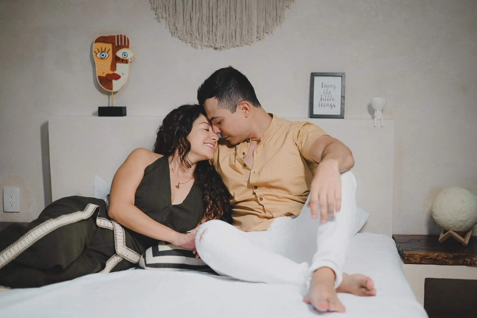 A laughing Latina woman with voluminous curly hair leans her head back in delight as her male partner nuzzles his face into her temple while…