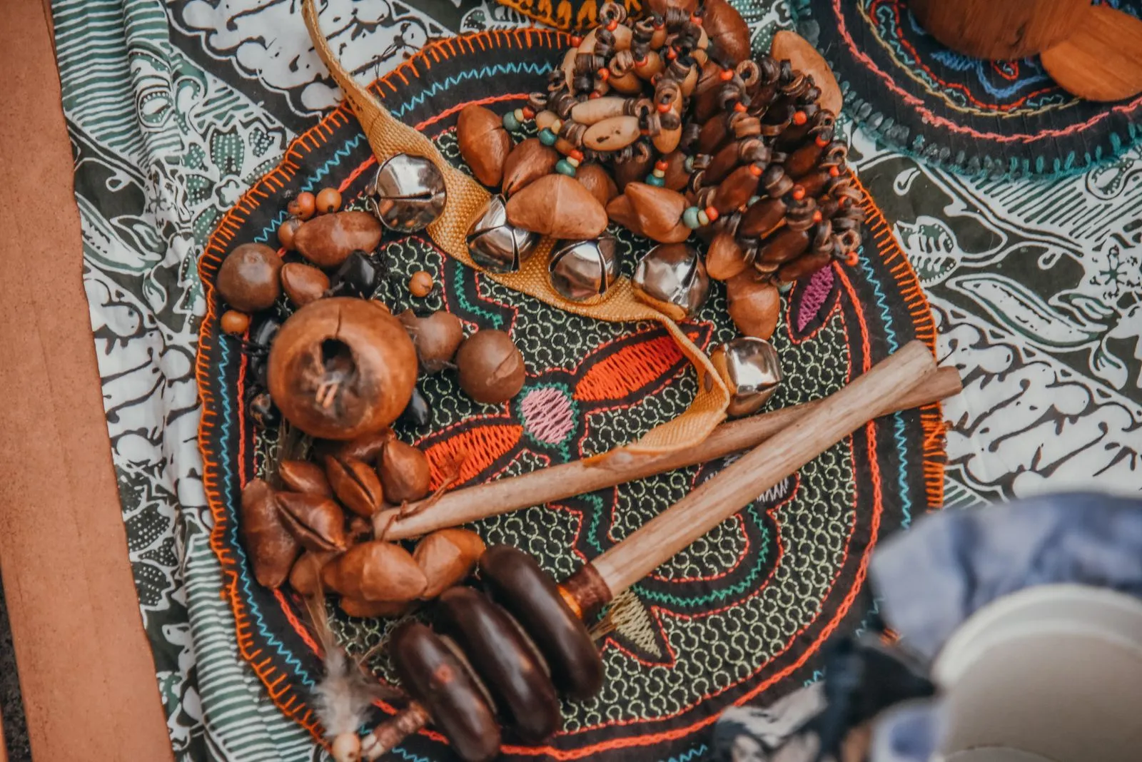 An overhead flat-lay of traditional Mesoamerican percussion instruments arranged on a hand-embroidered circular textile with orange and teal…
