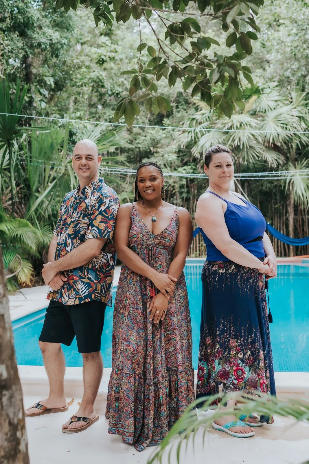 Three guests pose poolside in vibrant tropical attire, framed by dense jungle canopy and lush palms.