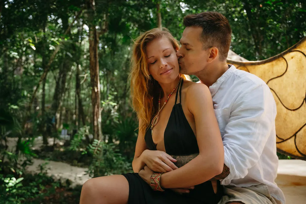 Two women embrace outdoors in neutral-toned linens, one with eyes closed in genuine connection. Soft natural light reveals a small beaded ankle chain detail and authentic affection
